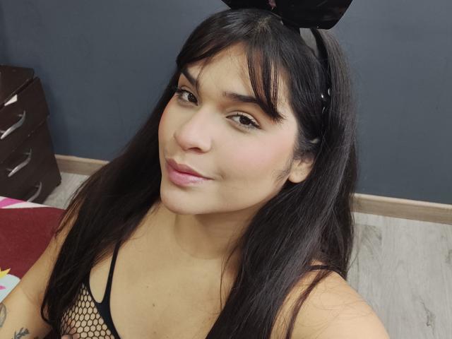 Antonela, avec de longs cheveux noirs, portant un haut en filet noir et des oreilles de lapin noires, souriant à la caméra, montrant de petites seins, des lèvres roses, une peau brun clair, un sol en bois, un mur sombre..