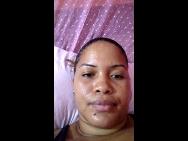 IrisI69 has a chubby face, brown skin, small nose, and a mole under her right lip. She's looking at the camera with a slight smile, wearing a black shirt.