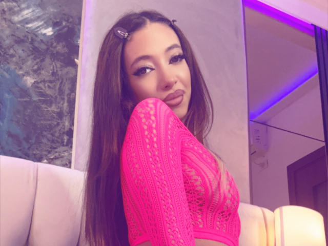 LaylaKrista with long brown hair, pink lace top, pouty lips, and hair clips, showing her tits, sitting on a white couch, purple lighting in the background.