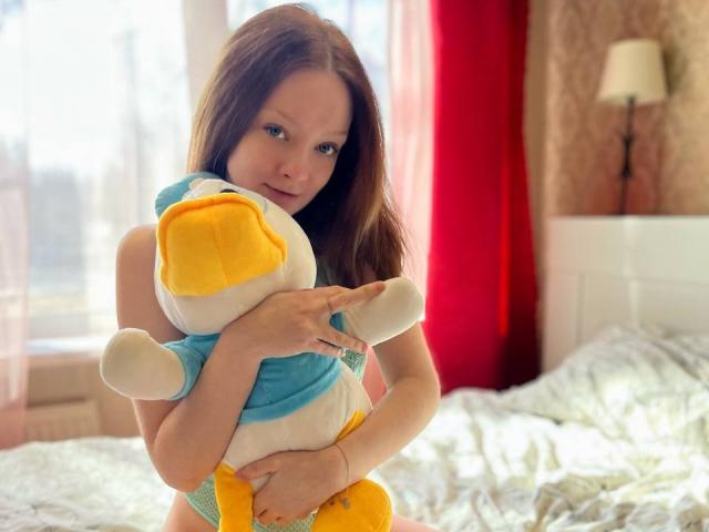 RadiantsLoves met bruin haar, die een geel en wit pluchen eend vasthoudt, gekleed in groene kantonderbroek, staand in een zonnige slaapkamer met rode gordijnen. Zachte glimlach, vriendelijke ogen..