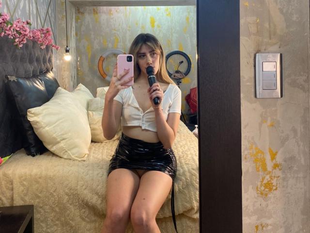 LivelyVanderWood with blonde hair, white crop top, and black shiny skirt, holding a microphone, sitting on a bed, taking a mirror selfie. Boobs visible under top.