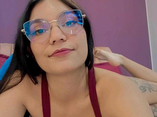 AriaLeenn with glasses, red halter top, black hair, tattoo on right shoulder, fair skin, pink lips, hand behind head, blue reflection in glasses, slight smile.