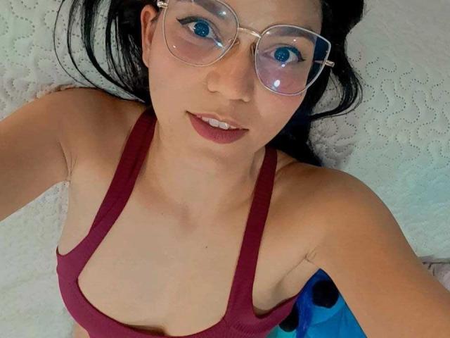AriaLeenn with glasses, blue eyes, and red lipstick, wearing a red tank top, showing her small tits and fair skin, lying on a white textured surface.