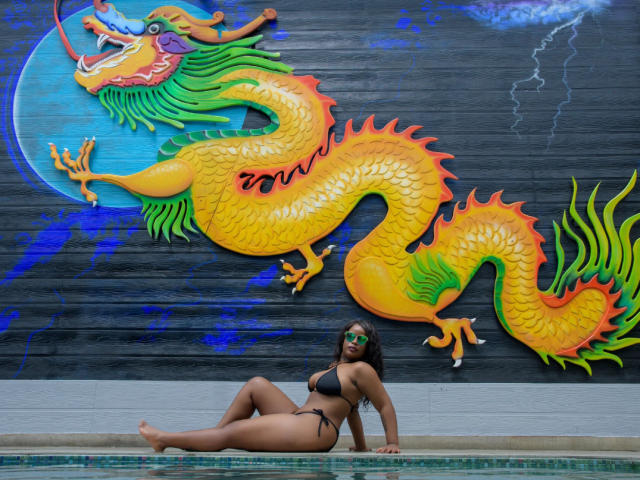 MadinsonLopez, with dark skin and green face mask, sits by the pool in a black bikini, showcasing her big ass and small tits, against a colorful dragon mural.