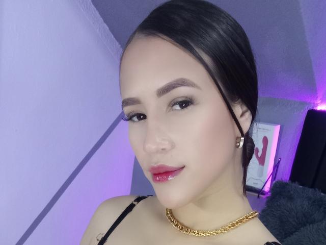 BeckaTheas with shiny pink lips, dark hair in a bun, gold chain necklace, and black strap, looking at camera with subtle smile, wearing black eyeliner, in a purple-lit room.