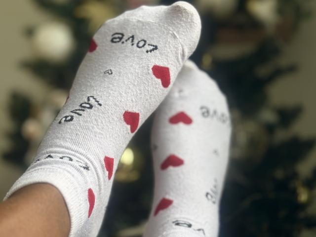 DeliceTropical's feet wearing white socks with red heart patterns and black text, one sock slightly lifted showing the bottom. Socks have a soft, slightly fuzzy texture.