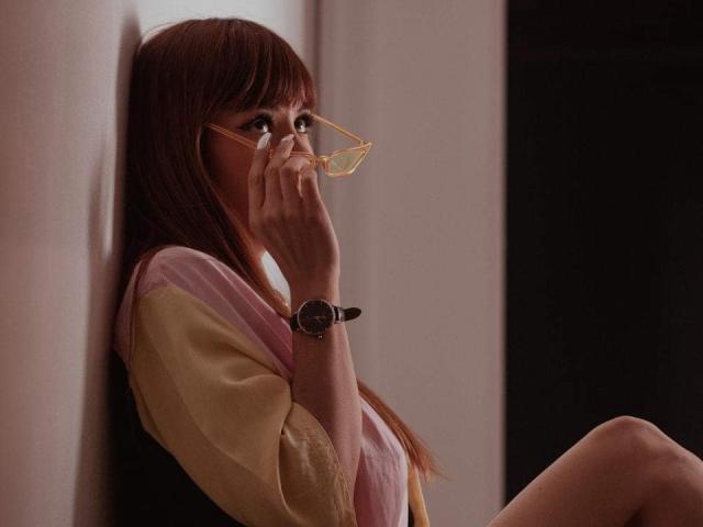 AriiannaHot with brown hair, wearing glasses, yellow and pink shirt, black vest, and wristwatch, sitting against wall, holding glasses, looking to side, in dim light.