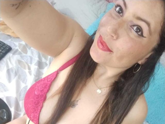 Vennus69 mit langen braunen Haaren, rotem Lippenstift, Nasenring und Creolen, trägt einen pinken Spitzen-BH, lächelt in die Kamera, zeigt ihre Brüste und liegt auf einem Bett..
