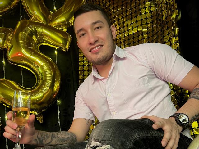 AironBlade with a smile, holding a glass of champagne, wearing a pink shirt, black jeans, and a watch, with a golden "5" balloon and gold glitter in the background.