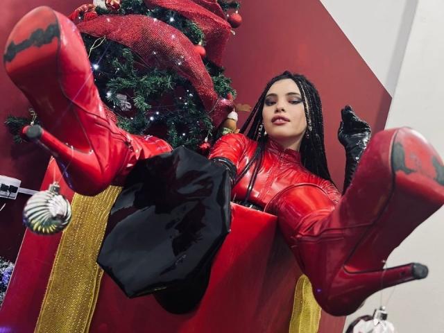 ElizaLovely in red latex outfit, black gloves, and red high heels, sitting in a red box with Christmas tree, black bag, and ornaments. Her legs are raised, showing soles.