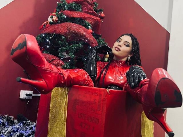ElizaLovely in red latex outfit, black gloves, and high heels, sitting in a giant gift box with legs up, next to a decorated Christmas tree.