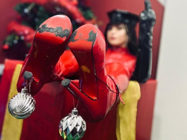 ElizaLovely in shiny red latex boots with black soles, wearing a matching latex suit and black gloves, legs up, Christmas ornaments hanging from her heels, blurred face.