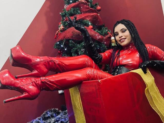 ElizaLovely with black braids, red latex boots, and gloves, sitting in a red gift box, legs up, smiling, next to a Christmas tree with red ribbons.