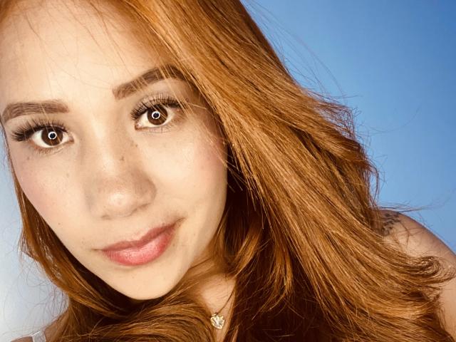 GraceHottieX has red hair, brown eyes, and a slight smile with pink lips. She has a tattoo on her right shoulder and wears a small necklace.
