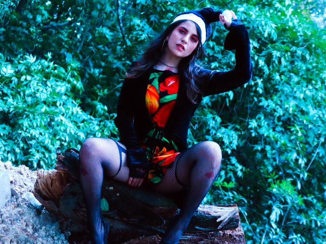 Araña Amy, con cabello oscuro, vestida con un atuendo negro con un estampado floral rojo-anaranjado, medias de rejilla y una diadema blanca, sentada sobre un tronco, con la mano en la cabeza, fondo de bosque..