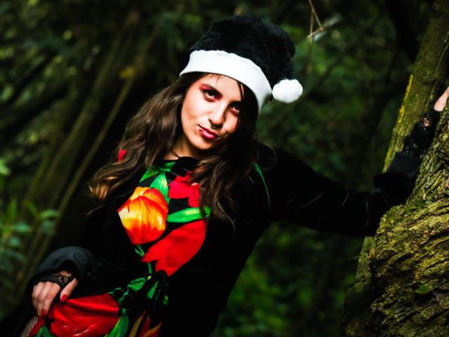 AmySpider with long brown hair, wearing a black Santa hat, black velvet dress with red and green Christmas ornaments, leaning against a mossy tree in a forest.