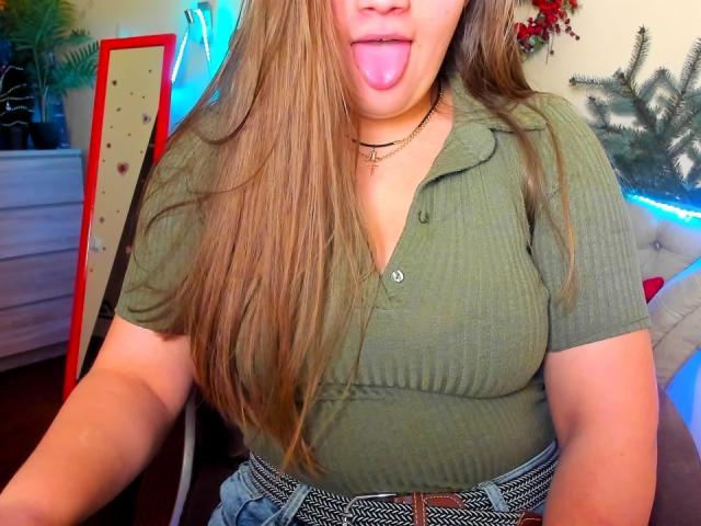 Madrinia with long brown hair, green ribbed shirt, and tongue out, wearing a belt with a brown buckle, standing in a room with blue neon light. Boobs visible.