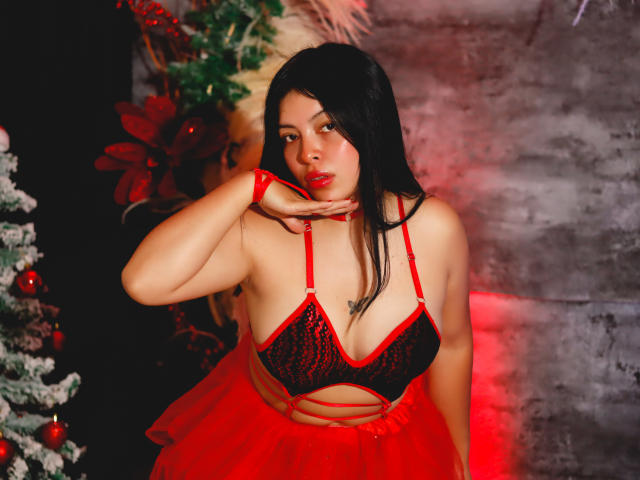 KateWelsin with dark hair, red and black lace bra, and red skirt, hand near chin, red wristband, red lipstick, and a slightly pouty expression, showing her tits.