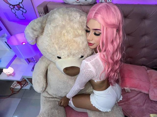 AlissLovely with pink hair, white crop top, and white shorts, snuggles against a giant fluffy teddy bear, showing her ass, in a pink-lit bedroom.