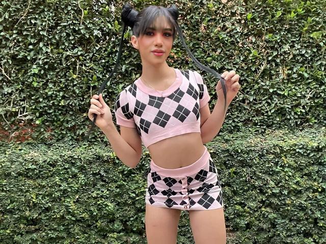 AlissLovely with black pigtails, pink and black diamond-patterned crop top and shorts, holding her pigtails, standing in front of a green hedge, showing her flat ass.