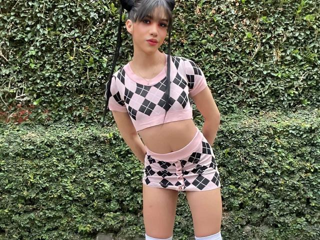 AlissLovely with pink and black diamond-patterned crop top and shorts, hands in pockets, standing against a green hedge. She's wearing thigh-high socks and has black hair in puffs.