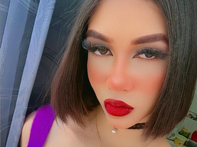 AlissLovely has a cute face with red lipstick, black choker, and purple top. She's got dark eyebrows, green eyes, and a septum piercing. Her brown bob is straight.