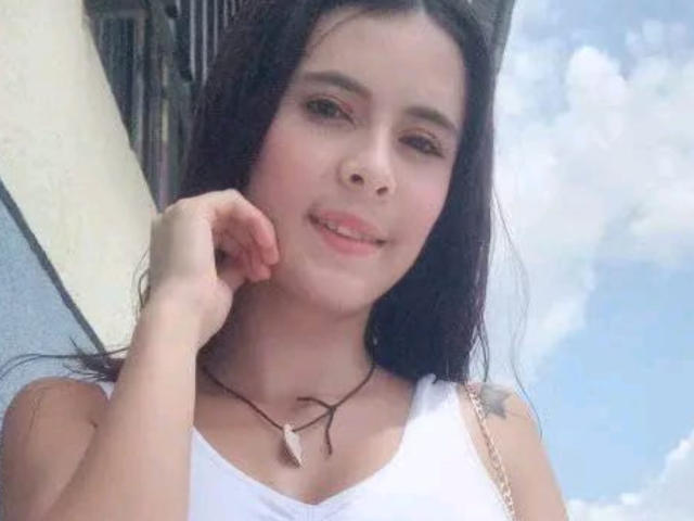 TiffanyBaez with long dark hair, white tank top, small tits, light skin, smiling, hand near face, necklace with wooden pendant, blue sky with clouds in background.