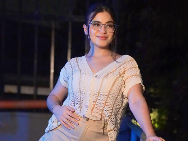 CamilaSofiia with glasses, white lace top, beige pants, hand on belly, smiling, dark hair in a bun, standing, dim lighting, soft blue and orange hues.