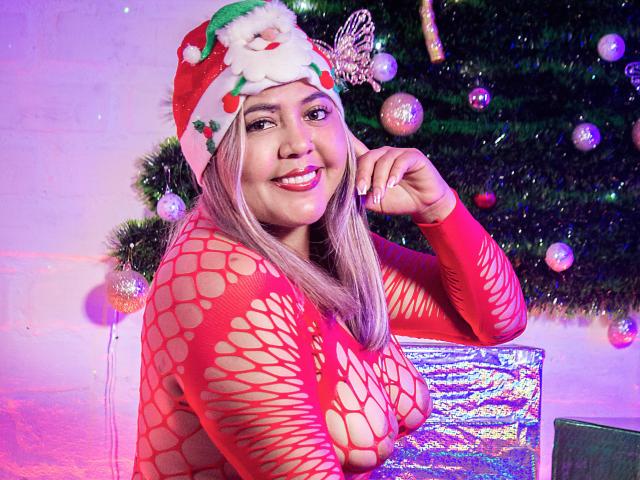 GraceJansen in a red, fishnet, long-sleeve top showing off her big, round tits, wearing a Santa hat with a white pom-pom, smiling, next to a Christmas tree.