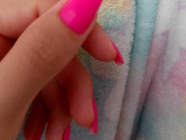 De roze, felroze gelakte nagels van KariCarter, afgebeeld tegen een gestructureerde handdoek. Haar nagels zijn lang en licht gebogen, met een gladde, verzorgde uitstraling..