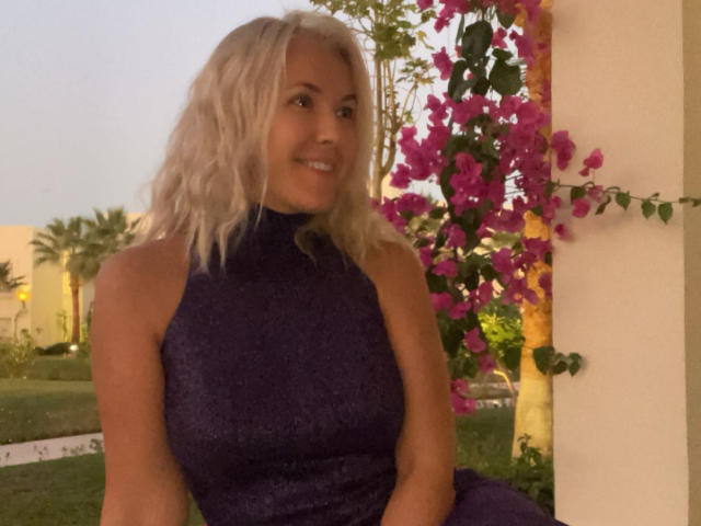 Misstyk with blonde wavy hair, smiling, wearing a sleeveless dark purple sparkly dress, standing by a wall with pink bougainvillea. Her tits are medium-sized, visible under the dress.