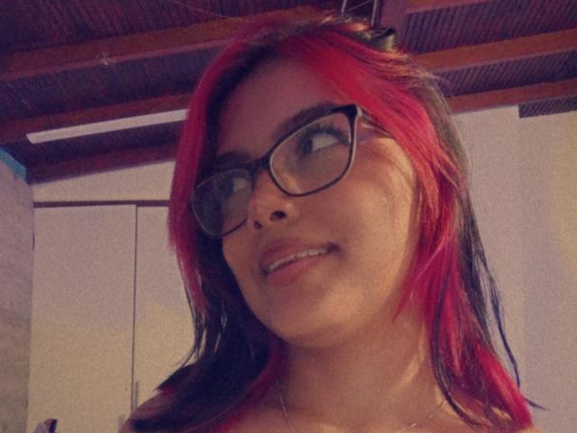 EmilyRoberts with red and black hair, wearing glasses, smiling slightly, showing teeth, in a selfie with wooden ceiling above.