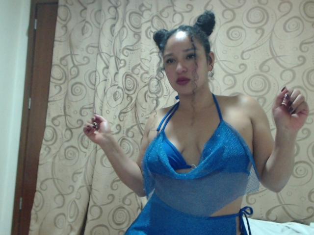 CarlaMuille with double buns, wearing a blue halter top that reveals her large tits and a matching skirt, posing with hands up, red lipstick, and dark nails.
