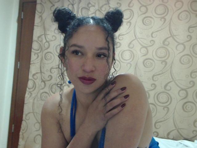 CarlaMuille with curly black hair in double buns, blue halter top, red lipstick, and dark red nails, sitting with knee up, looking at camera with slightly parted lips.