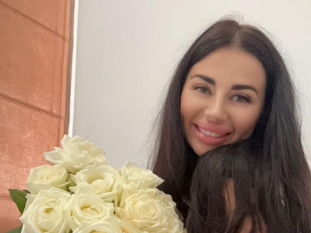 CuddliesElis with long dark brown hair, smiling, holding a bouquet of white roses, has pink lipstick, and brown eyes, in a selfie.