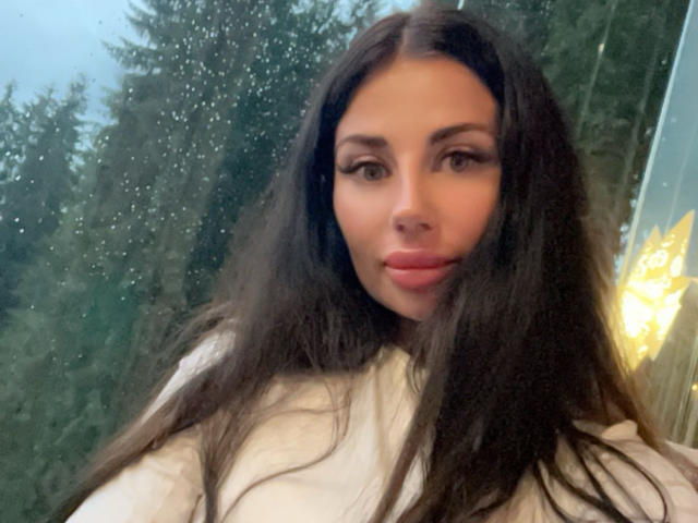 CuddliesElis with long brown hair, full lips, and brown eyes, wearing a white top, looking at the camera with a slight smile, standing in front of a window.