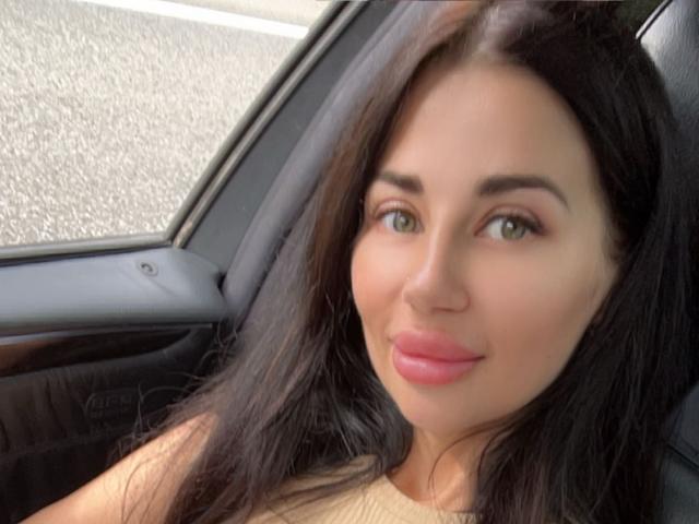 CuddliesElis has medium tits, green eyes, pink lipstick, and long dark hair, sitting in a car with a subtle smile.