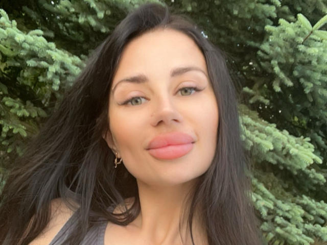 CuddliesElis has puffy pink lips, green eyes, and long black hair. She's wearing gold hoop earrings and looking slightly to the side with a subtle smile.