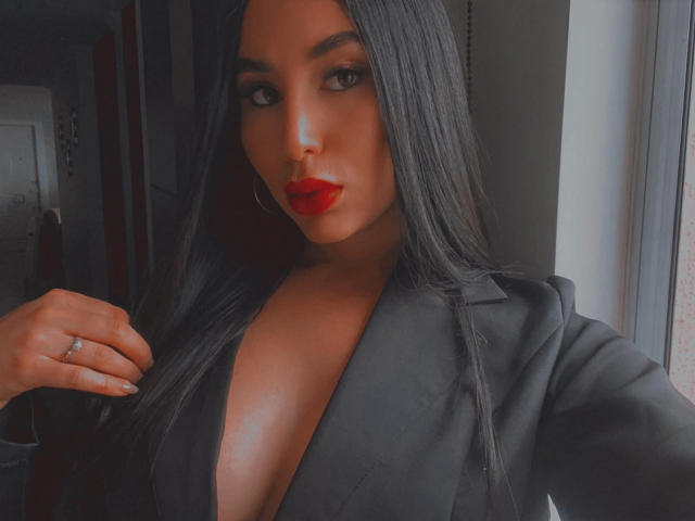 AgathaRuiz with long black hair, red lipstick, and a deep black V-neck shirt showing her big tits. She's holding her hair, wearing a ring, near a window.