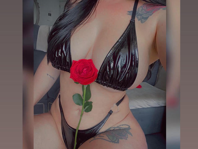 AgathaRuiz with black shiny bikini, red rose between her tits, tattoos on arms and thigh, long black hair, holding rose in front of her crotch, in a car.