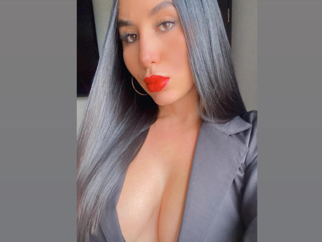 AgathaRuiz, avec ses longs cheveux noirs, son rouge à lèvres rouge et son chemisier en satin noir à décolleté plongeant qui révèle ses grandes seins. Elle porte des boucles d'oreilles dorées et a un regard confiant..