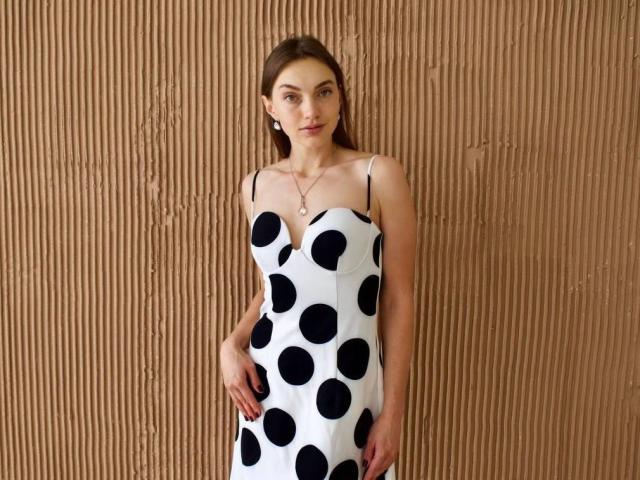 HoneyRusset wearing a white dress with black polka dots, standing against a textured brown wall, has small tits, a slim figure, and brown hair, looking at the camera.
