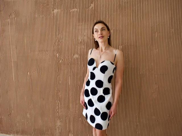 HoneyRusset stands against a brown corrugated wall wearing a white dress with black polka dots, showing her slim, pale body and small tits, with a neutral expression.
