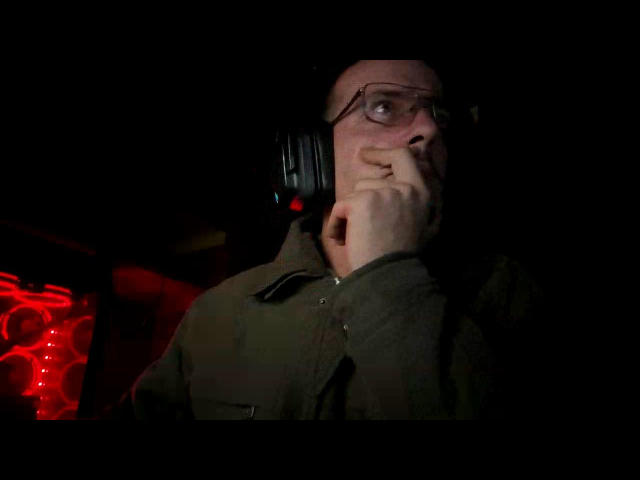 MasterDonJuan with glasses, wearing headphones, has a finger to his lips, looking up with a slightly surprised or curious expression, wearing a green shirt, in dim lighting.