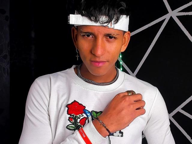 ThiagoFabian with brown skin, black curly hair, white headband, white shirt, red rose graphic, green earrings, chain necklace, ring, bracelet, looking serious, medium shot, black background.