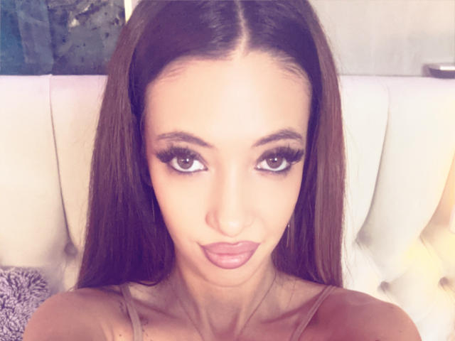 LaylaKrista has long brown hair, big brown eyes with long lashes, and full pink lips. She's wearing a tank top and sitting on a white tufted couch.
