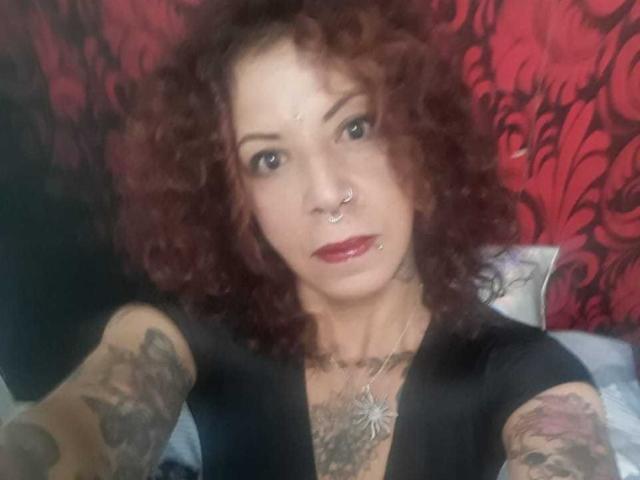 Axxy, com cabelo ruivo cacheado, piercing no nariz e batom vermelho, exibindo tatuagens nos braços e no peito, vestindo uma blusa preta, olhando para a câmera..