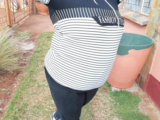 MatureBussty's striped shirt and black pants, showing a chubby ass, with a green trash can and red house in the background. Grass and dirt path on the side.