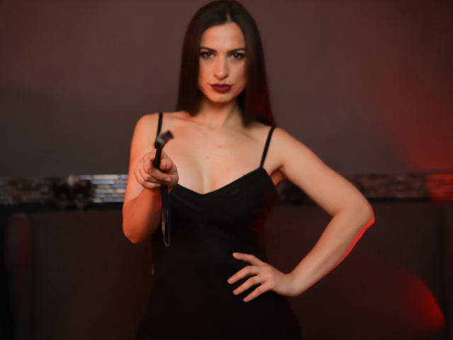 QueenSerenne, com longos cabelos castanhos, vestindo um vestido preto, segurando um chicote preto, com a mão na cintura, iluminação vermelha, expressão séria, plano médio, magra com seios pequenos, confiante..