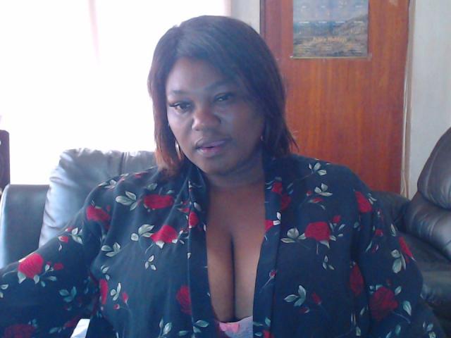 MatureBussty with dark skin and brown hair, wearing a black floral robe with red roses, showing big tits in a pink bra, sitting on a leather couch.